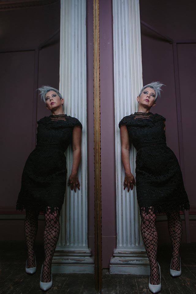 A woman wearing a black dress and leaning against a white wooden column, the whole reflected in a large mirror.