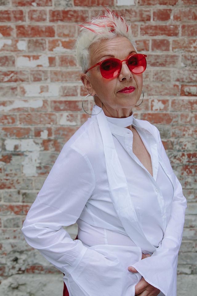 Older woman with short, spikey white hair and large red glasses wearing a white blouse, behind her a plain brick wall.