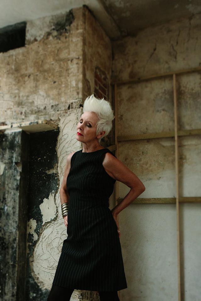Woman with white spiky hair wearing a black pinstriped dress and standing in a derelict room.