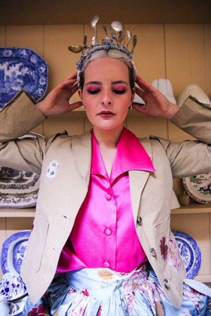 Photo of woman in a light jacket over a vivid pink top. She is wearing a head dress comprising old teaspoons and is posing with her hands held high, by her ears.