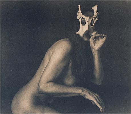 Photo: head and torso portrait of a woman, side-on and naked against a dark background. In one hand she is holding to her face part of an animal skeleton, through which she stares back at the viewer.