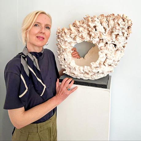 Photo of the artist standing, one hand resting on an abstract sculpture supported by a plinth. 