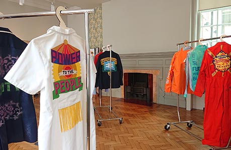 Photograph of colourful garments decorated with text, on clothes rails in a Regency interior