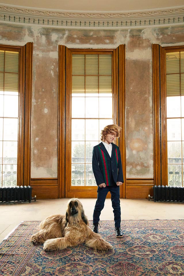 Photo of a boy wearing jeans and a blue jacket with red and green stripes. He is standing in an undecorated room with large windows in the background, an Afghan Hound is sitting by his feet.