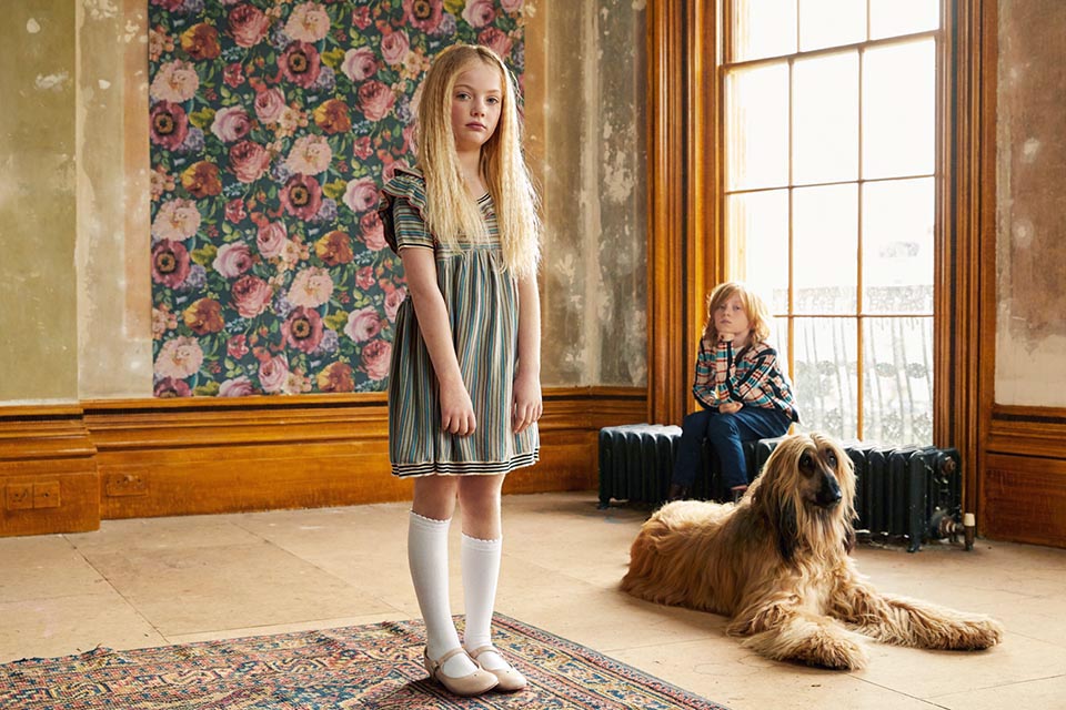 Photo of a little girl standing in a large undecorated room, behind her sits a little boy, at her feet is an Afghan Hound.