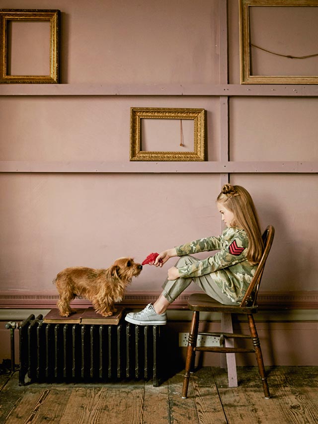 Photo of a little girl sat in a chair and pointing a red water pistol at a small dog.
