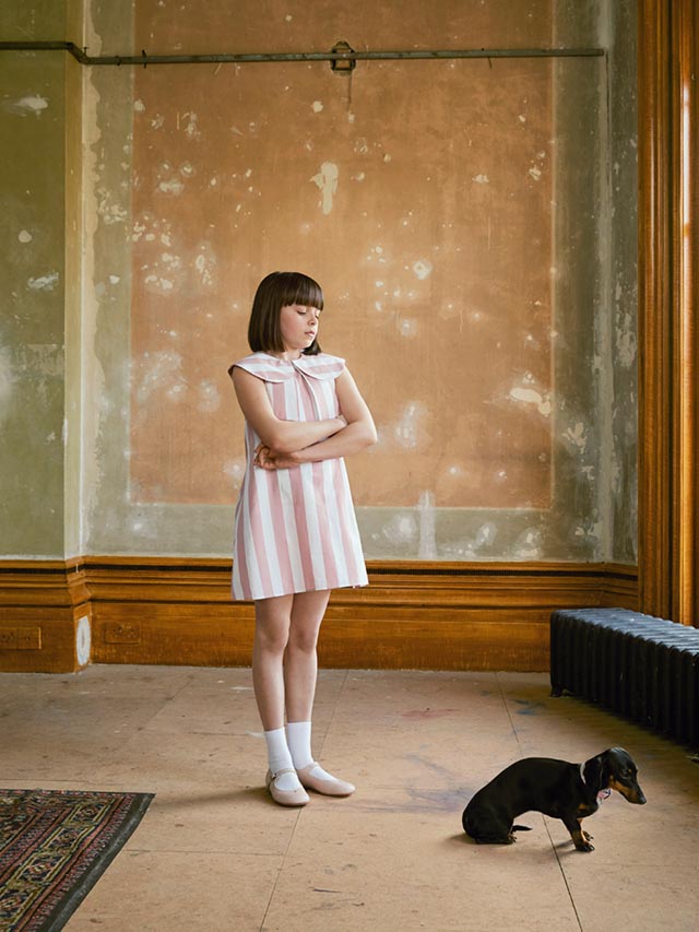 Photo of a girl wearing a white and pink striped dress, standing in an undecorated room and looking down at a small Daschund. 