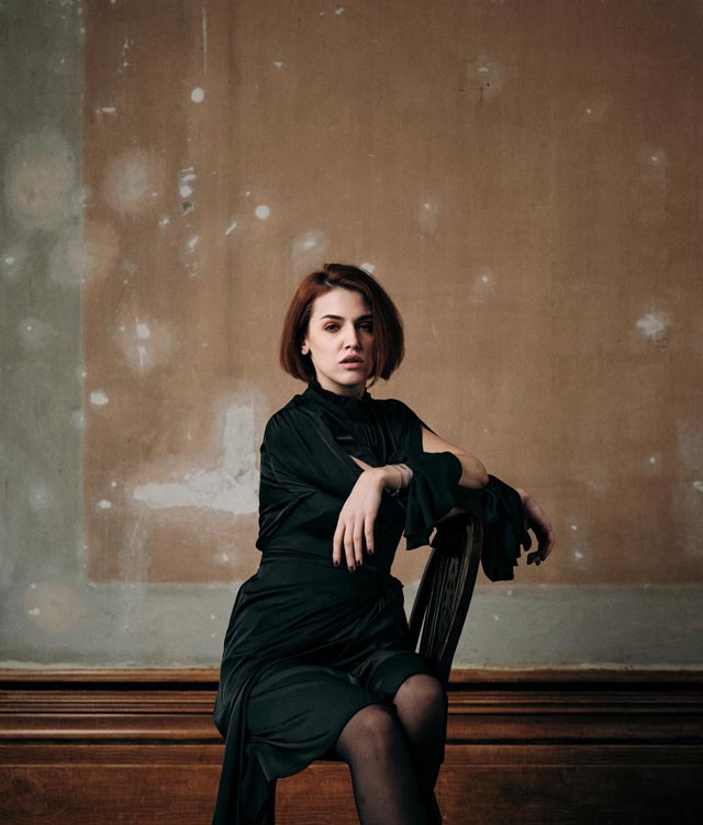 Photo of a young woman wearing a dark, knee-length dress, sitting on a wooden chair, with a bare plaster wall in the background.