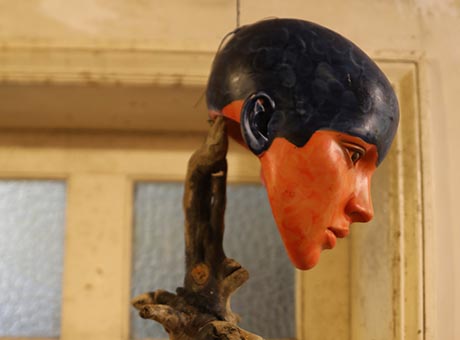 Photo showing a mannequin head, painted orange and black, supported by a tree branch