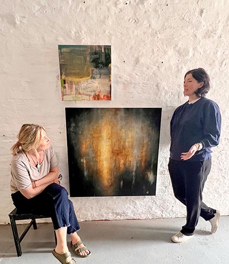 The artists Kate Scott, seated, and Abigail Bowen, standing, discuss two artworks on the wall between them