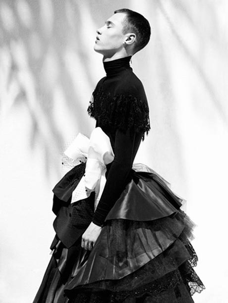 Black and white photo of a man in profile, he is wearing a black top and layered skirt with a large white bow at the waist.