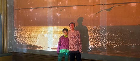 Photo of a woman and a man standing in front of a wall, onto which is projected a sunset over the sea