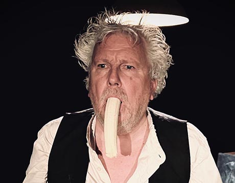 Photo of a dishevelled man with thinning white hair. A banana is hanging out of his mouth.