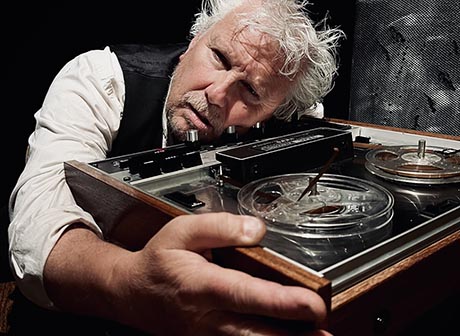 Photo of a dishevelled man with thinning white hair and rolled-up sleeves. He is seated and resting his head on an old tape recorder while embracing it with an outstretched arm.