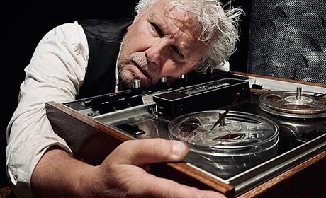 Photo of a dishevelled man with thinning white hair and rolled-up sleeves. He is seated and resting his head on an old tape recorder while embracing it with an outstretched arm.