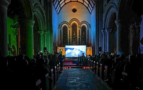 Photo of church interior looking down the centre aisle towards the chancel. It is dark, peole are sat in the pews and a large screen shows a projected image of a landscape.