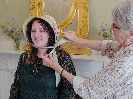 Photo of a woman trying on a bonnet helped by another woman who ties the ribbon to hold it on