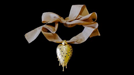 A gold heart-shaped amulet on a silk ribbon. It is decorated with small black spots and is dripping gold 