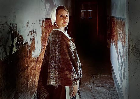 Photo of a woman standing in a dark corridor. She is wearing white cloth bonnet and a dark woolen cloak with an apron beneath.