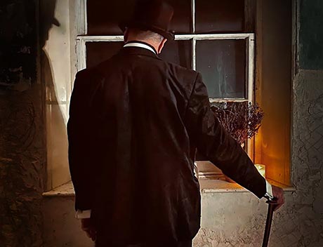 Photo of a man with his back to the camera. He is in a room with bare plaster wall and is wearing dark suit, top hat, and his right hand rests on a cane.