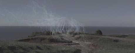 Landscape photo, taken looking across a headland and out to sea. There is a fence and whispy, smoke-like trails in the air.