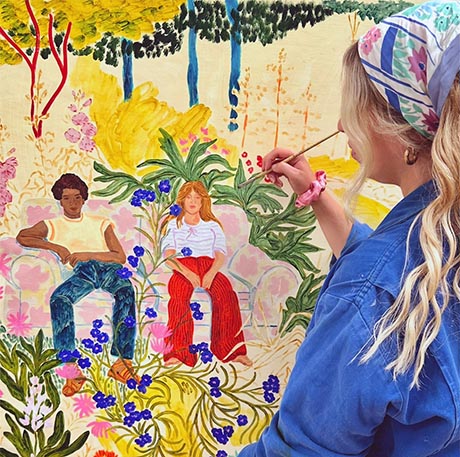 Photo of an artist at work. She is wearing a blue overall and headscarf and we look over her shoulder as she paints two figures seated in a colourful garden landscape.
