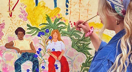 Photo of an artist at work. She is wearing a blue overall and headscarf and we look over her shoulder as she paints two figures seated in a colourful garden landscape.