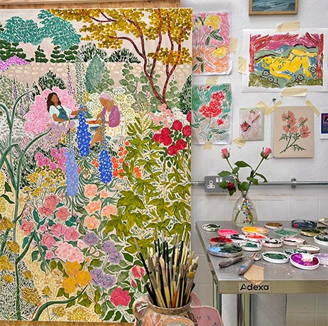Photo of an artist's studio. A colourful garden landscape is propped up, in the foregrond a jug full of brushes, to the right a table is covered in coloured paint lids, in the background there are sketches taped to the wall.
